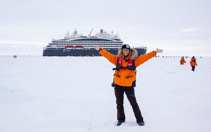 Female traveler and AdventureSmith specialist wears orange parka with fur lined hood, arms spread out, posing in snowy landscape of North Pole in front of Le Commandant Charcot ship.