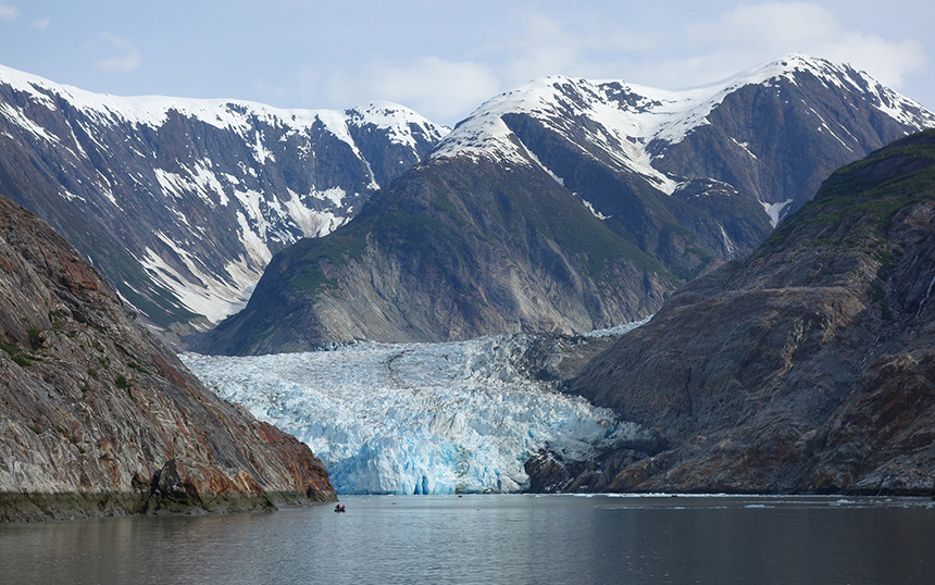 A small skiff seen in the water near a large glacier with snow-covered mountains behind it in Tracy Arm in Alaska's Inside Passage.