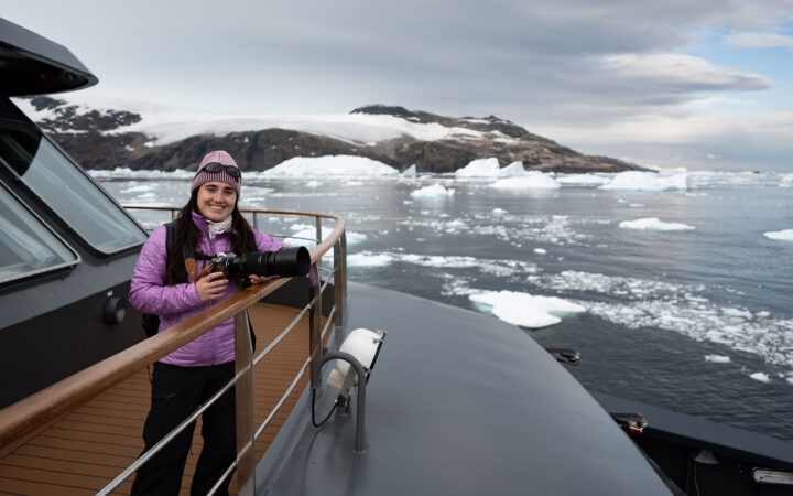 A female traveler in a purple jacket stands on the bow of a ship in Antarctica holding a camera leaning on the rail