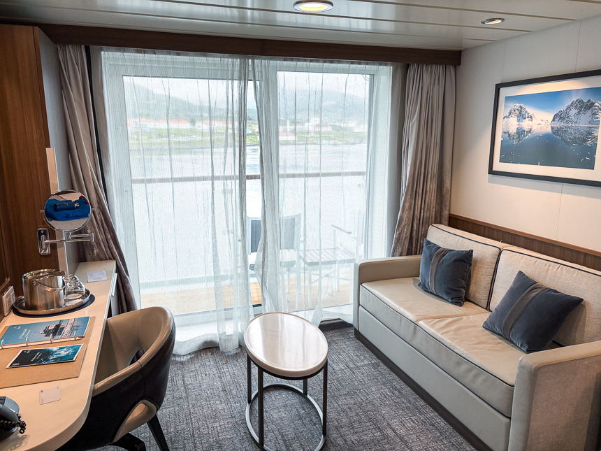 Guest room aboard Sylvia Earle with floor-to-ceiling windows, balcony, couch, and vanity area with tan and blue design