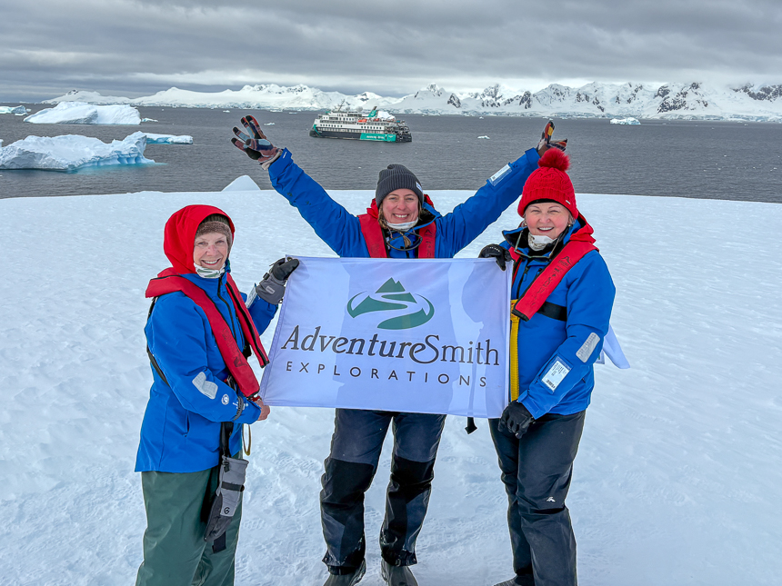 Three adventurers standing on land in antarctica with one women with her hands in the air and an AdventureSmith flag between the 2 other women, with Sylvia Earle off in the distance.