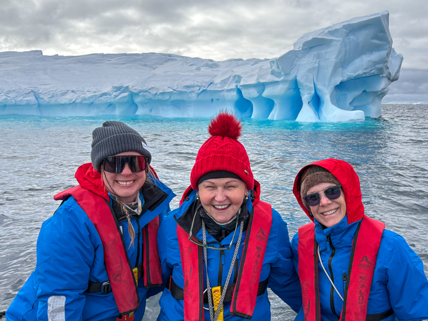 Three adventurers dressed in red and blue are smiling in Antarctica in front of a large iceberg on a sunny day 