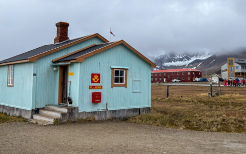 Teal house sits among green & gold arctic tundra backed by tall peaks on a cloudy day in Ny Alesund, Svalbard.