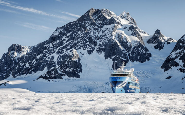 Blue & teal small expedition ship with pointed bow sits among ice beside dark snowy peaks on a sunny day in Svalbard.