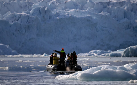 Zodiac boat with Svalbard circumnavigation travelers moves among floating iceberg bits beside tall white glacier on a sunny day.