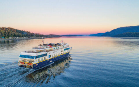 Small expedition ship with dark blue hull & white upper decks cruises in glassy water at sunset in the AK & BC inside passage.