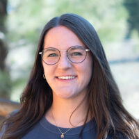 Portrait of Brie Gorter. She's wearing a navy blue shirt and glasses. Pine trees and a wooden fence can be seen in the background.
