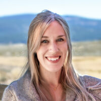 Portrait of Adventure Coordinator Olivia Williams. Blonde female wearing grey sweater leaning on wooden fence