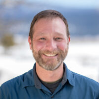 adventuresmith explorations director of sales and operations justin massoni wearing a blue shirt smiling on a sunny day with snow and mountains in the background