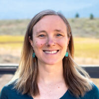 AdventureSmith Content Manager Mary Montalbano with blonde hair & teal shirt stands in front of a wooden fence & golden field.