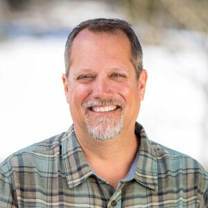 A portrait of AdventureSmith founder Todd Smith smiling looking at camera in a plaid shirt with fence behind him