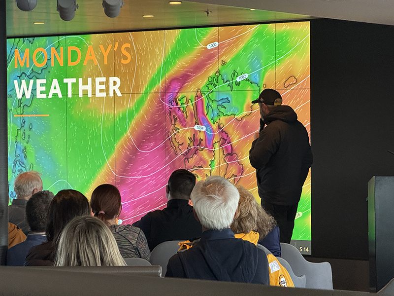 A guide shows a big screen of a weather map showing Monday's Weather writing and color overlays showing wind