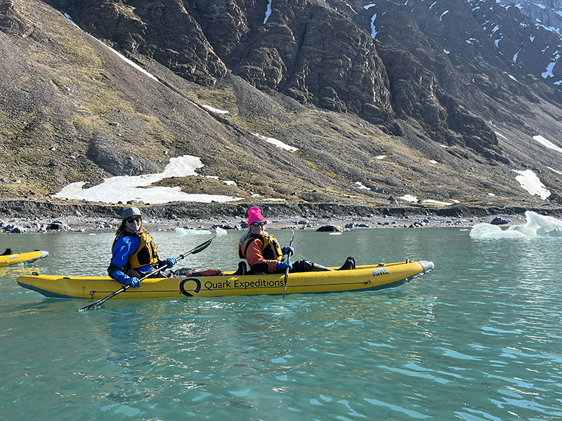 Two females in a yellow kayak smile with a brown mountainous shoreline behind them dotted with snow