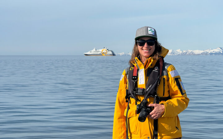 Woman in yellow jacket holding camera on Arctic shore, expedition ship Ultramarine and snowy peaks behind under blue skies.