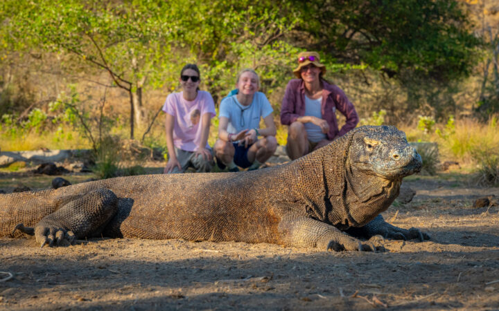 Mom & 2 daughters in summer clothing pose behind a large komodo dragon with scaly, tan skin on the Sailing Indonesia: Komodo & Beyond cruise.