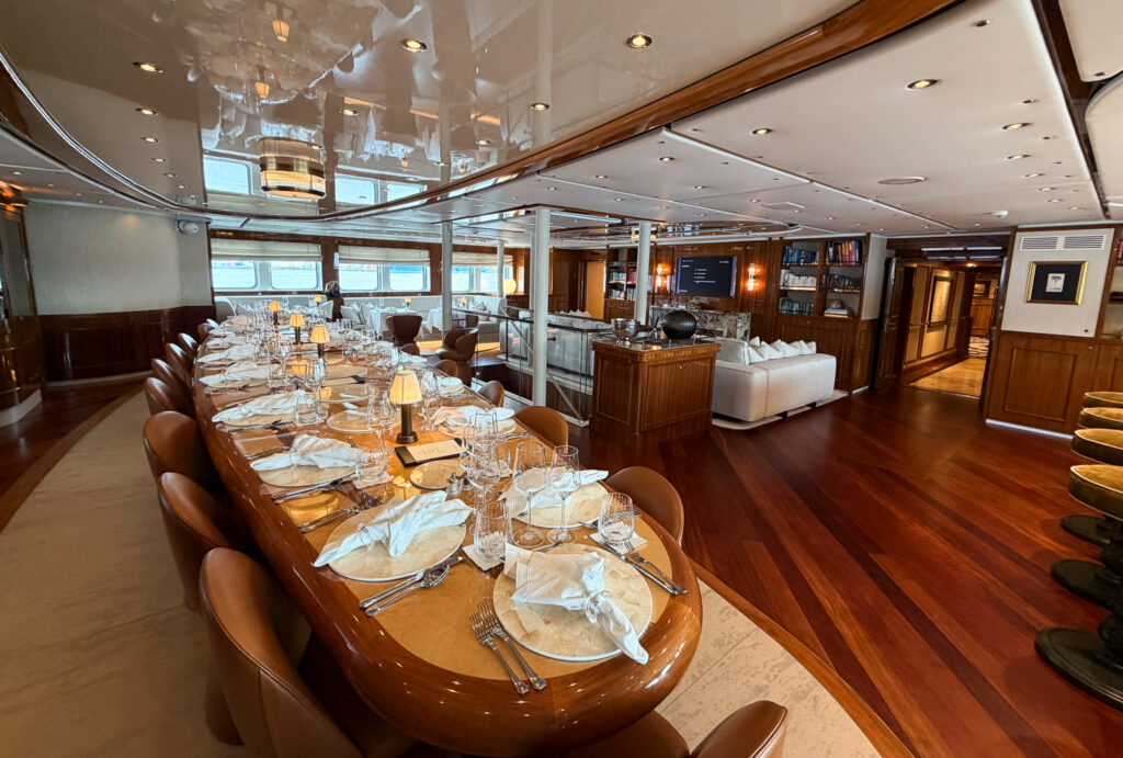 Salon aboard Aqua Lares luxury yacht, one long table for guests to sit for meals, surrounded in rich wood and white appointments. 