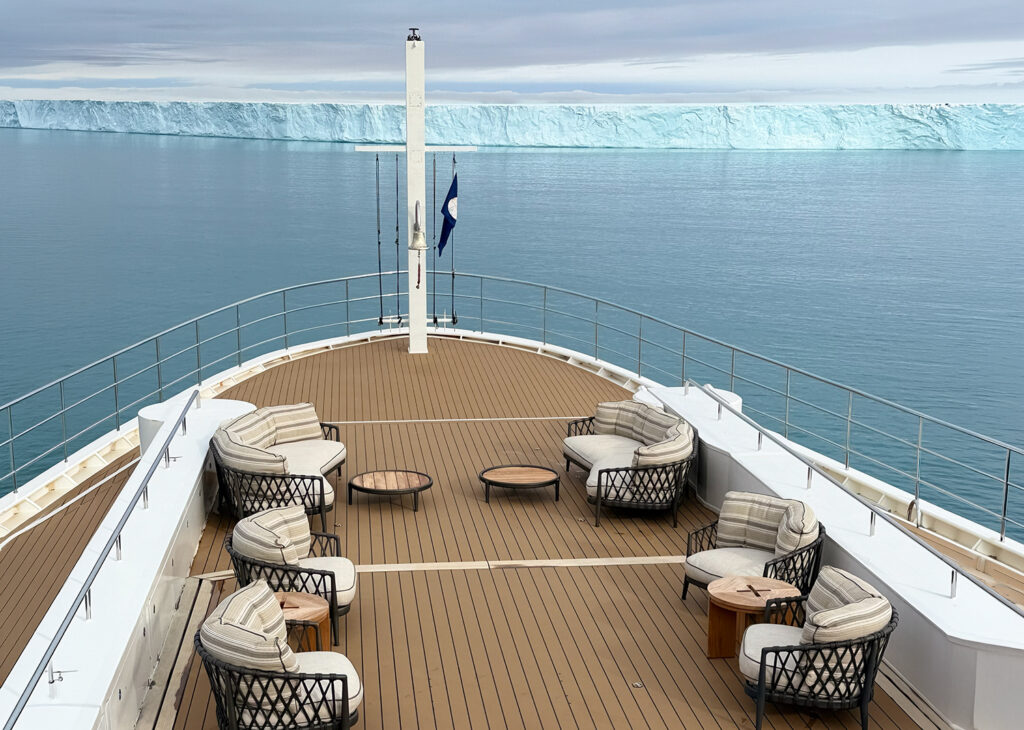 The bow of Aqua Lares yacht, with deck furniture, anchored in front of a massive wall of ice at Bråsvellbreen glacier. 