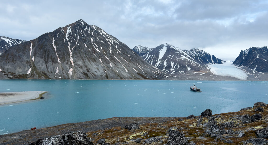 From shore, you can see Aqua Lares yacht anchored in quiet cove of teal water surrounded by jagged mountains with veins of snow. 
