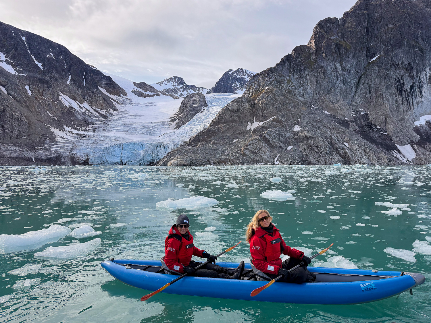 Two female travelers in inflatable double blue kayak wearing red dry suits and life jackets, paddle in front of glacier and among ice. 