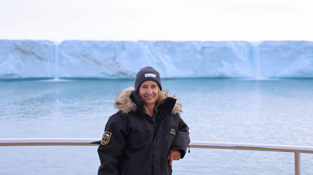 AdventureSmith expert wearing Aqua Lares black parka with fur hood stands on bow of ship, behind her is a massive wall of ice at Bråsvellbreen glacier