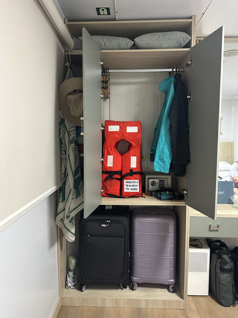 Small beige closet with open doors & 2 suitcases beneath aboard Galapagos Angel.