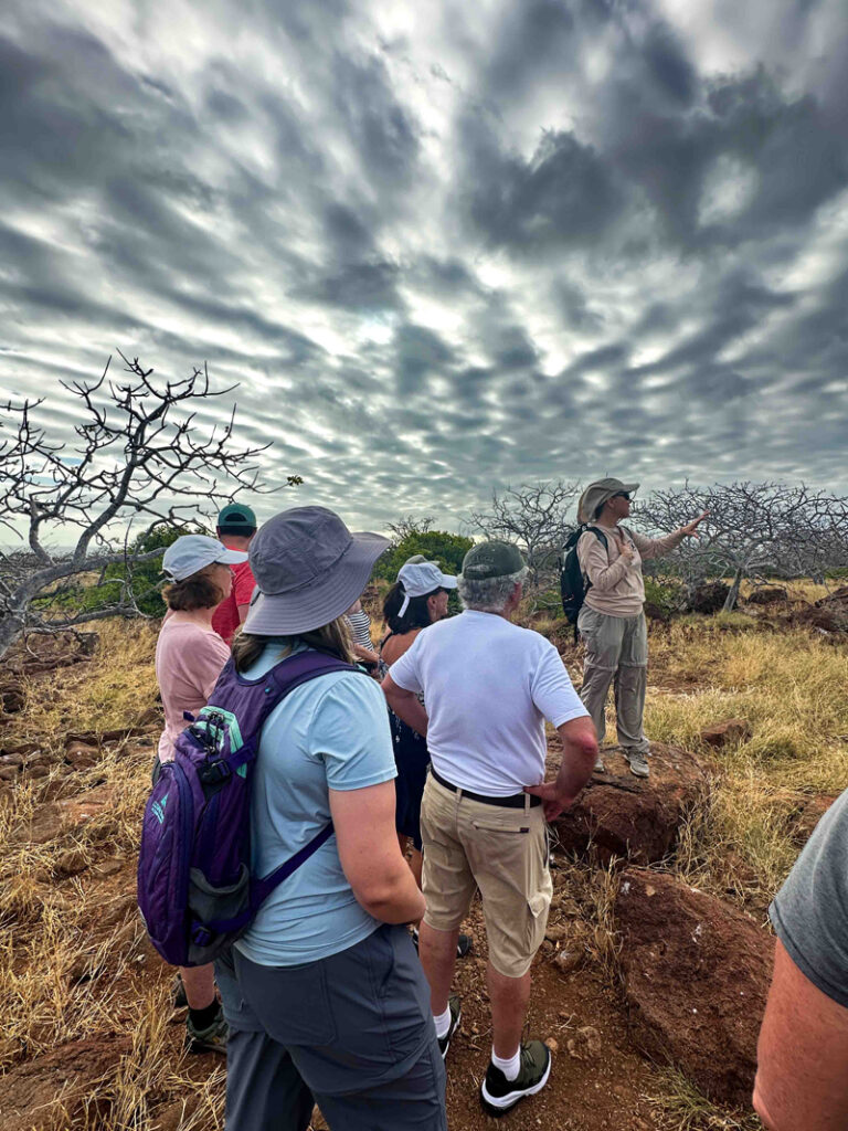 Group of Galapagos travelers stands & listens to a guide under cloudy sky.