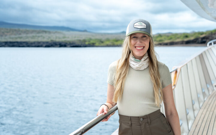 Woman in beige & olive shorts & t-shirt stands on deck of the Galapagos Angel yacht on a sunny day.