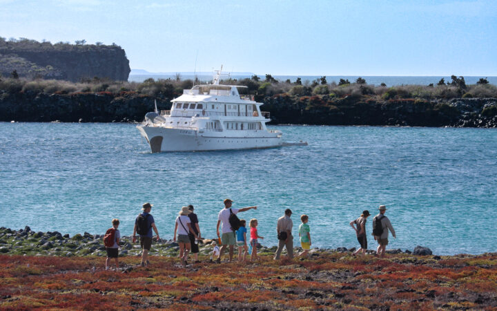 Galapagos cruise deals aboard Coral I ship that floats offshore as family group of travelers walk on colorful trail.