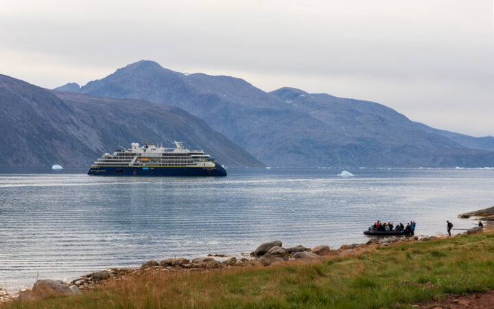 Small ship with dark blue hull & white upper decks sits offshore from grassy coast where Zodiac of Greenland travelers are arriving.