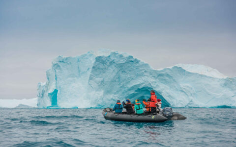 Zodiac boat with south Greenland Exploration cruise guests in colorful coats sits in front of large blue iceberg in calm sea.
