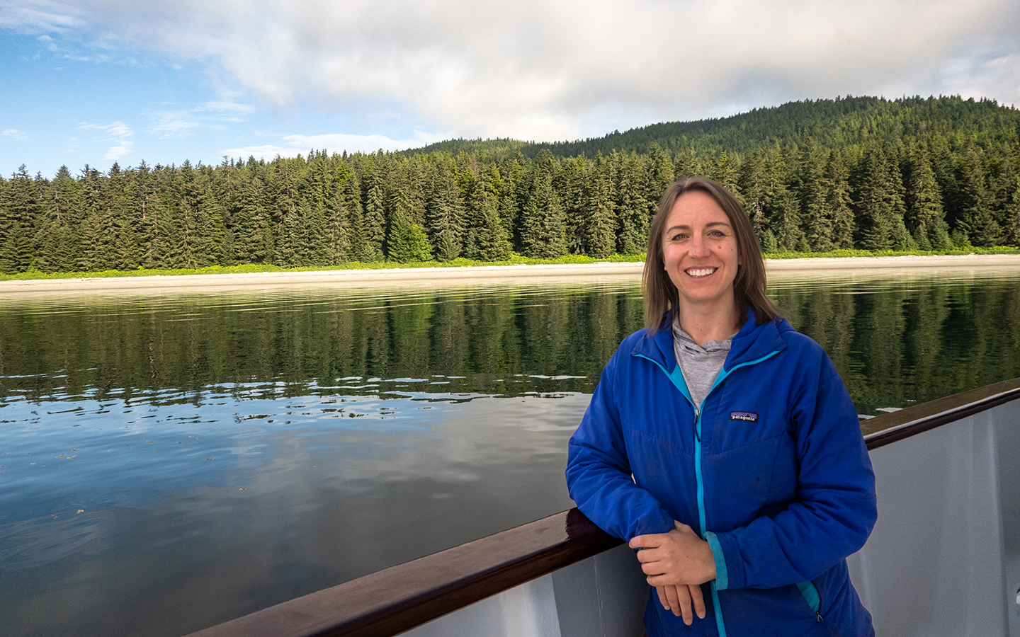 Female Alaska expert in royal blue jacket leans over ship railing with forested alaska shoreline behind her