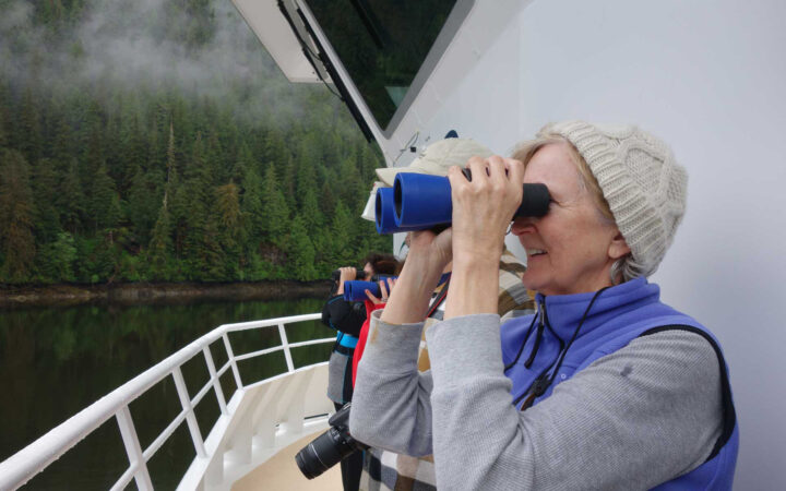 Female traveler stands on bow of Alaska small ship and looks through blue binoculars