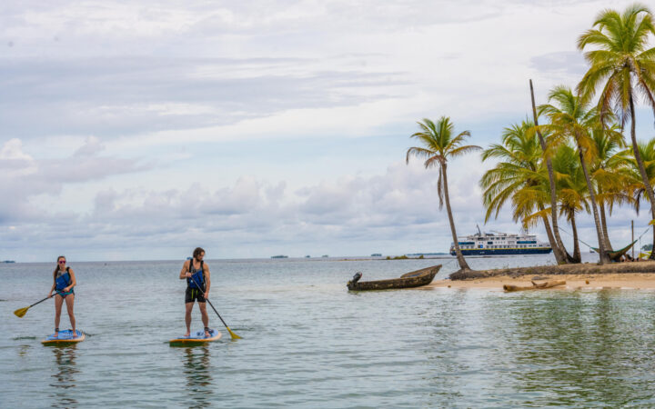 Man & woman paddle stand-up paddlebaords by shore of tiny island with white-sand beach & palm trees as ship sits nearby in Panama.