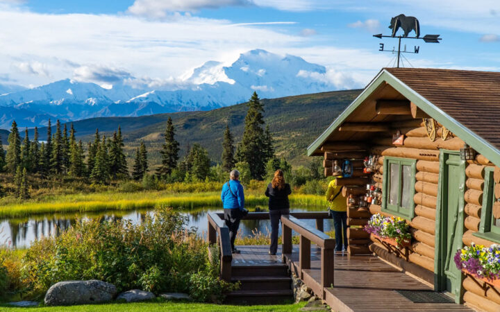 Two travelers stand on deck of Camp Denali wooden lodge taking in the sweeping views of a lake, wilderness and clear sight of Denali mountain in the distance.