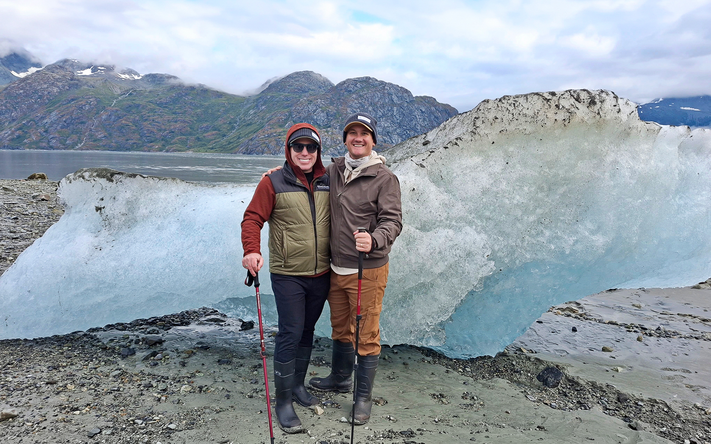 Two men pose on shore in Glacier Bay National Park with hiking sticks and a massive beached iceberg behind them