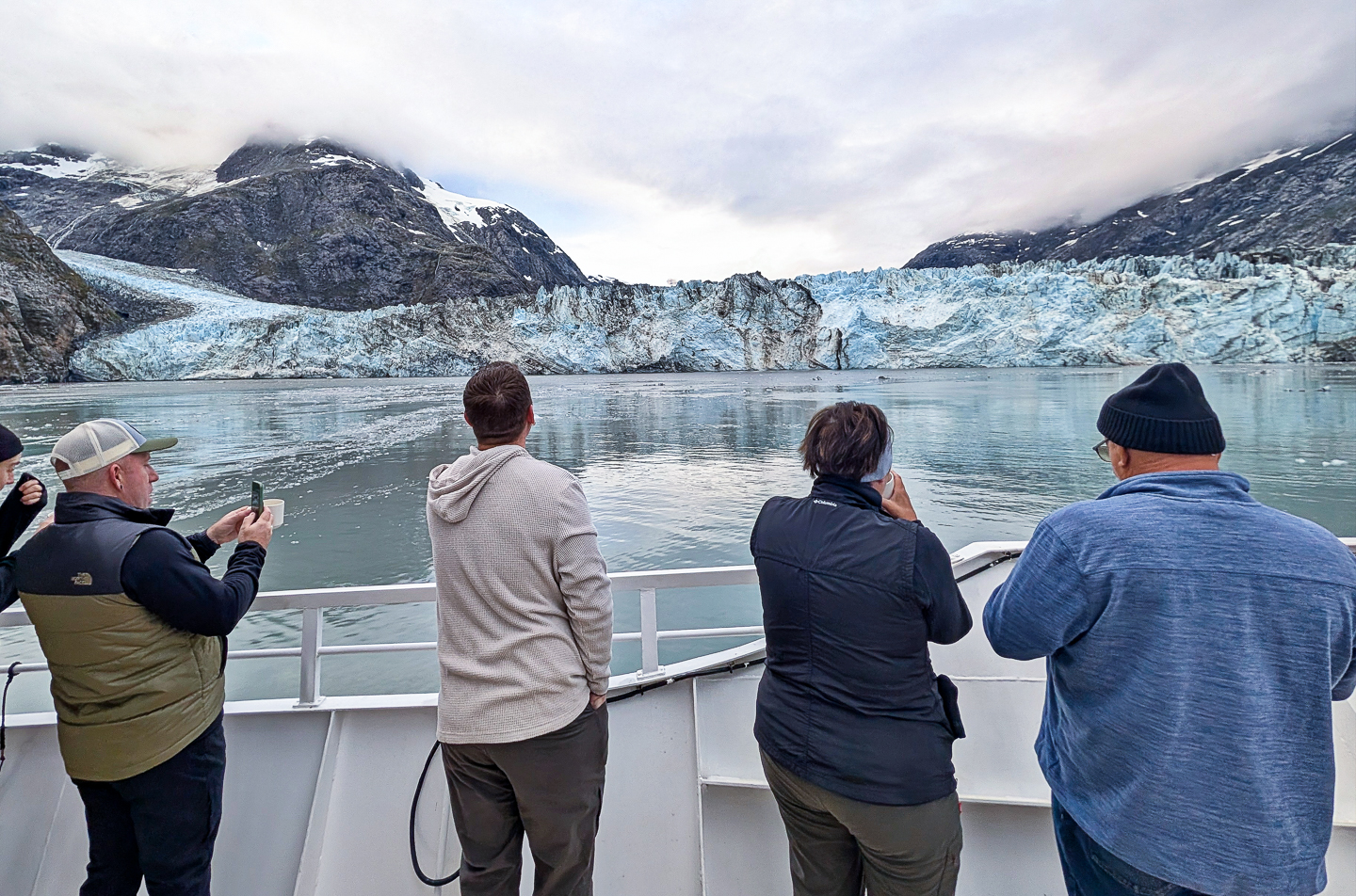 In Glacier Bay National Park, travelers gather on bow of ship taking photos of the teal blue glacier in front of them
