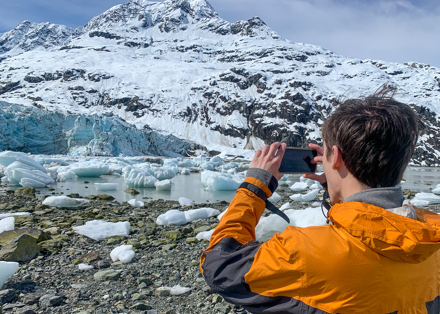 Boy in orange parka stands on shore among field ice bergs and photographs a glacier and snow capped mountains