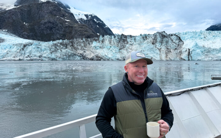 Man wears hat and vest and holds white cup of coffee as he stands on bow of small ship that is anchored close to the face of a glacier in Glacier Bay National Park.
