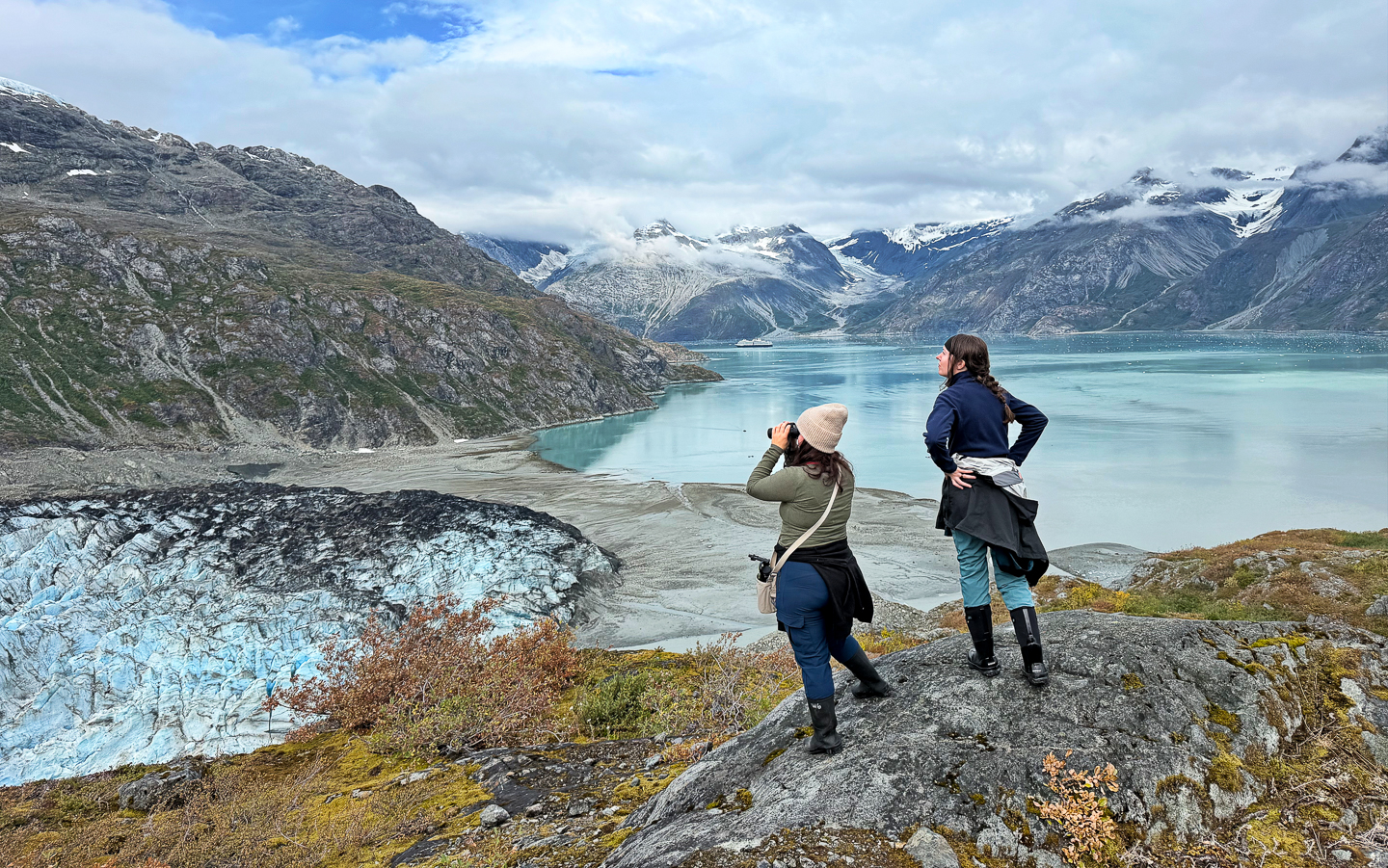 Two female travelers stand on the edge of a vista point high above Glacier Bay and look at sweeping views of teal water and a glacier. 