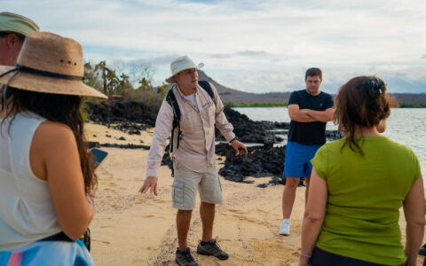 Galapagos guide in khaki stands in circle of travelers on beach & explains what guests are seeing on an Eminence cruise.